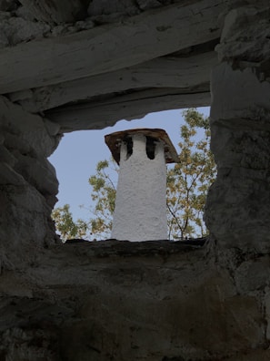 A stone chimney with a rustic appearance is viewed through an opening framed by rough, textured stone walls. The backdrop features green foliage against a clear blue sky.