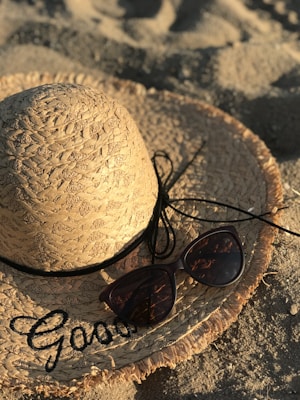 A straw hat with intricate weaving patterns lies on sandy ground, adorned with black lettering and black cord. Beside it, a pair of dark-tinted sunglasses casts a shadow on the hat.