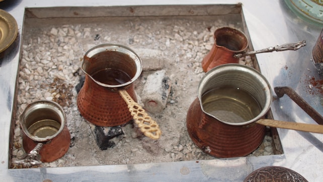 Several traditional copper pots, known as cezves, are arranged on a bed of hot sand, used for brewing coffee. The pots have long handles, and some contain liquid. The scene appears to be an outdoor setup, possibly for serving traditional Turkish coffee.