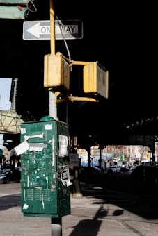 A weathered traffic signal post with yellow traffic lights and a one-way sign is installed above a large green utility box covered in torn papers and graffiti. The scene is set under an overpass, casting shadows on the street, with the less busy road and parked cars visible in the background.