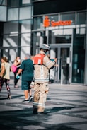 A person in firefighter gear stands on the street while interacting with a small device. There are people walking around casually with a modern building and a bank entrance in the background.