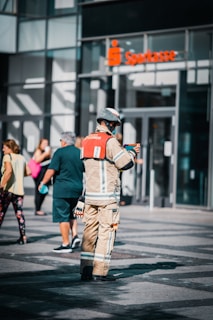 A person in firefighter gear stands on the street while interacting with a small device. There are people walking around casually with a modern building and a bank entrance in the background.