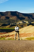 A person stands on a rocky terrain with a camera and tripod, capturing the unique geological landscape of striped hills and distant dark mountains under a clear blue sky.
