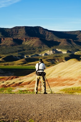 A person stands on a rocky terrain with a camera and tripod, capturing the unique geological landscape of striped hills and distant dark mountains under a clear blue sky.