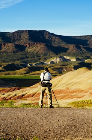 A person stands on a rocky terrain with a camera and tripod, capturing the unique geological landscape of striped hills and distant dark mountains under a clear blue sky.