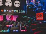A gaming setup featuring a Samsung monitor displaying graphics of stormtrooper helmets and various software icons. Below the monitor, there is a colorful RGB-backlit keyboard. On the right side, a Redragon mousepad is visible next to a gaming mouse lit with neon colors. In the background, parts of a computer with a Gigabyte graphics card and RGB lighting are seen.