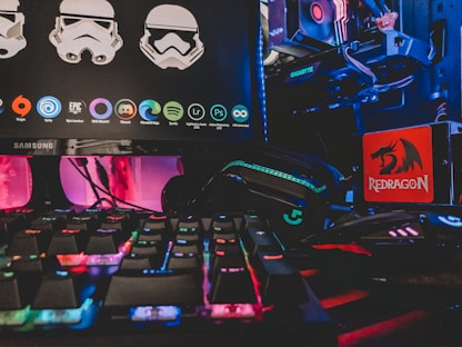 A gaming setup featuring a Samsung monitor displaying graphics of stormtrooper helmets and various software icons. Below the monitor, there is a colorful RGB-backlit keyboard. On the right side, a Redragon mousepad is visible next to a gaming mouse lit with neon colors. In the background, parts of a computer with a Gigabyte graphics card and RGB lighting are seen.