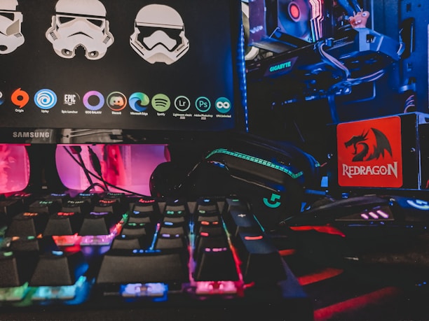 A gaming setup featuring a Samsung monitor displaying graphics of stormtrooper helmets and various software icons. Below the monitor, there is a colorful RGB-backlit keyboard. On the right side, a Redragon mousepad is visible next to a gaming mouse lit with neon colors. In the background, parts of a computer with a Gigabyte graphics card and RGB lighting are seen.