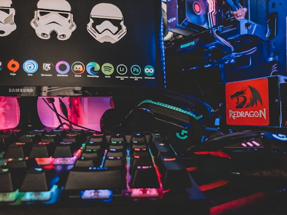 A gaming setup featuring a Samsung monitor displaying graphics of stormtrooper helmets and various software icons. Below the monitor, there is a colorful RGB-backlit keyboard. On the right side, a Redragon mousepad is visible next to a gaming mouse lit with neon colors. In the background, parts of a computer with a Gigabyte graphics card and RGB lighting are seen.