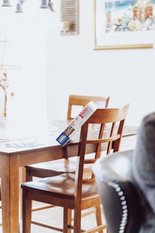 A wooden dining table and chair set is positioned in a well-lit room, with a puzzle box placed on one of the chairs. The puzzle box is leaning against the table, and puzzle pieces are scattered on the table surface. A bright, possibly natural light comes from the left, enhancing the cozy atmosphere. On the wall, there is a framed picture featuring a coastal scene.