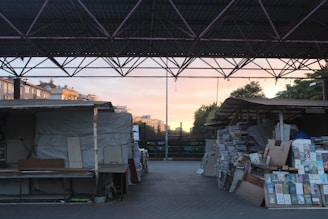A covered market area with stacks of books and wooden stalls on either side. The roof structure is composed of metal beams, and the sunlight from the setting sun illuminates the background, creating a contrast between the shadowy foreground and the lit buildings and sky.