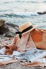 A serene beach scene features a straw hat with a black band, a woven tote bag, and a pair of brown sandals laid out on a striped towel. An open magazine rests casually on the towel. The background shows the ocean with gentle waves lapping against the rocks.