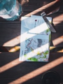 A table features a topographic map with detailed contours and vibrant colors. A knife with a wooden handle is placed nearby, next to a plastic water bottle. Sunlight casts dramatic shadows across the wooden surface, creating an adventurous atmosphere.