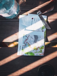 A table features a topographic map with detailed contours and vibrant colors. A knife with a wooden handle is placed nearby, next to a plastic water bottle. Sunlight casts dramatic shadows across the wooden surface, creating an adventurous atmosphere.
