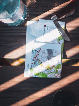 A table features a topographic map with detailed contours and vibrant colors. A knife with a wooden handle is placed nearby, next to a plastic water bottle. Sunlight casts dramatic shadows across the wooden surface, creating an adventurous atmosphere.