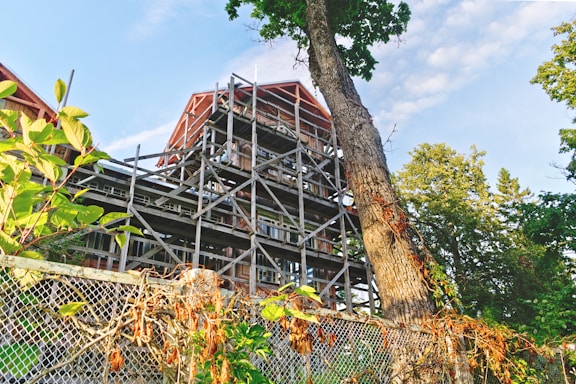 A partially constructed or scaffolded building structure is visible, surrounded by lush green trees and vines. The frame of the building appears to be made of metal or wood, with distinct diagonal beams. Some vegetation is growing around a mesh fence in the foreground, adding a natural and untamed feel to the scene.