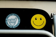 Two stickers are displayed on a black surface. The first is a circular sticker with the words 'NO WORRIES' and 'ONE LIFE LIVE IT' surrounding a hand sign. The second sticker is a bright yellow smiley face with black eyes and a curved smile.