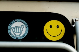 Two stickers are displayed on a black surface. The first is a circular sticker with the words 'NO WORRIES' and 'ONE LIFE LIVE IT' surrounding a hand sign. The second sticker is a bright yellow smiley face with black eyes and a curved smile.