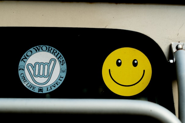 Two stickers are displayed on a black surface. The first is a circular sticker with the words 'NO WORRIES' and 'ONE LIFE LIVE IT' surrounding a hand sign. The second sticker is a bright yellow smiley face with black eyes and a curved smile.