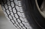 A close-up view of a vehicle tire showcasing its detailed tread pattern. The grooves and ridges are clearly visible, indicating a robust design, suitable for traction and durability. The tire sidewall has some branding visible, and the surface is slightly worn, suggesting it has been in use.