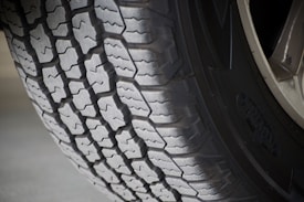 A close-up view of a vehicle tire showcasing its detailed tread pattern. The grooves and ridges are clearly visible, indicating a robust design, suitable for traction and durability. The tire sidewall has some branding visible, and the surface is slightly worn, suggesting it has been in use.