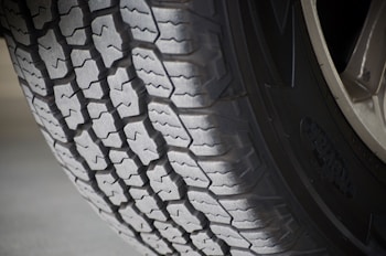 A close-up view of a vehicle tire showcasing its detailed tread pattern. The grooves and ridges are clearly visible, indicating a robust design, suitable for traction and durability. The tire sidewall has some branding visible, and the surface is slightly worn, suggesting it has been in use.