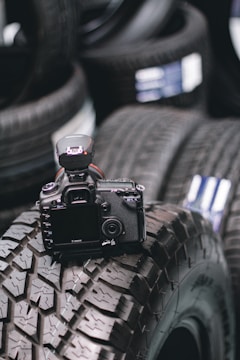 A DSLR camera is placed on top of a stack of rugged, all-terrain tires. The scene highlights the juxtaposition between technology and industrial themes, with the camera's lens and buttons visible. The focus is primarily on the camera, with the tires providing a textured background.
