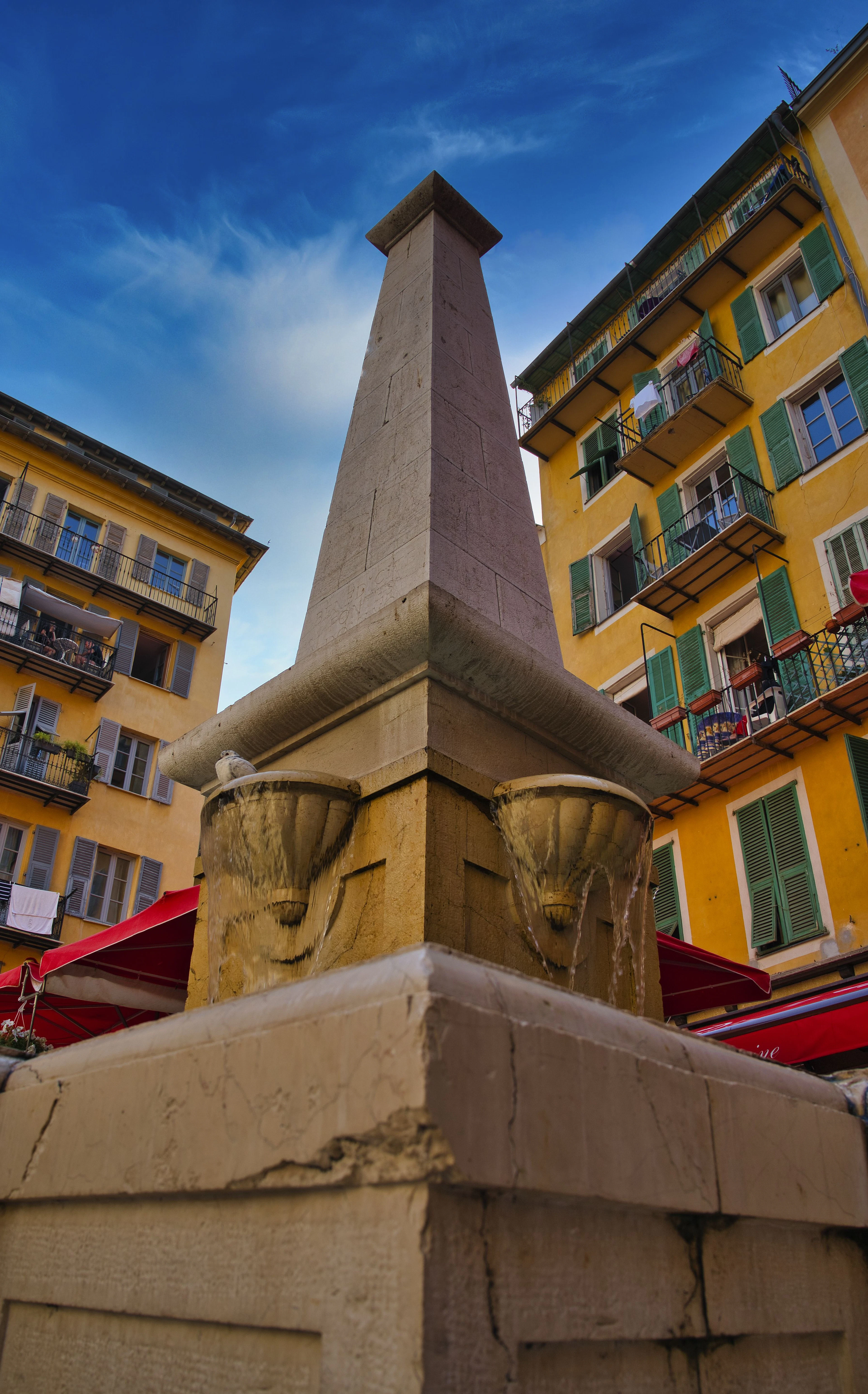 A stone obelisk fountain stands amid the yellow, green-shuttered buildings of Nice's old town.