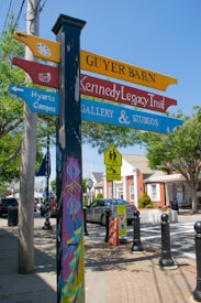 A colorful directional signpost is situated on a street corner, with various signs pointing to locations such as 'Guyer Barn', 'Kennedy Legacy Trail', and 'Hyarts Campus'. The post is decorated with vibrant geometric patterns. In the background, there's a crosswalk, vehicles, and buildings, indicating a small town setting. Trees line the street, and a school crossing sign is visible.