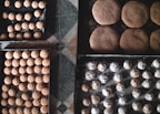 Trays filled with freshly baked cookies arranged neatly on a checkered floor. The cookies vary in shape and size, some dusted with powdered sugar and others having a round, dome-like form. The textures of the cookies are visually appealing, with some showing cracks on their surfaces.