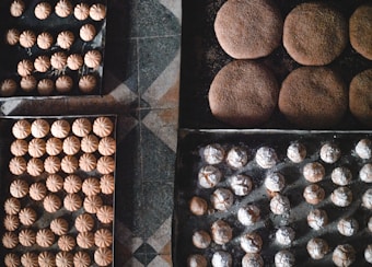 Trays filled with freshly baked cookies arranged neatly on a checkered floor. The cookies vary in shape and size, some dusted with powdered sugar and others having a round, dome-like form. The textures of the cookies are visually appealing, with some showing cracks on their surfaces.