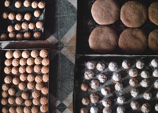 Trays filled with freshly baked cookies arranged neatly on a checkered floor. The cookies vary in shape and size, some dusted with powdered sugar and others having a round, dome-like form. The textures of the cookies are visually appealing, with some showing cracks on their surfaces.