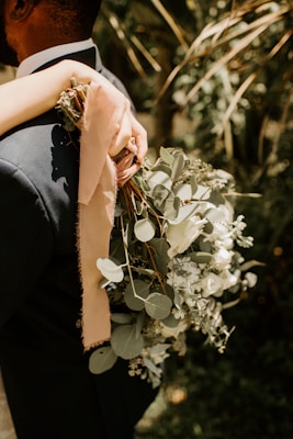 A person wearing a dark suit is holding a bouquet of greenery and white flowers, wrapped with a delicate pink ribbon. There is an arm wrapped around the person's shoulder, suggesting an intimate moment. The background features blurred greenery, enhancing the focus on the bouquet.
