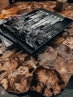 A wooden table with a unique, irregular pattern on its surface, featuring a large black-and-white photography book titled 'NEW YORK' depicting a city street scene. The book is partially overlaid with another book or magazine in the corner. A small wooden block is visible in the background.