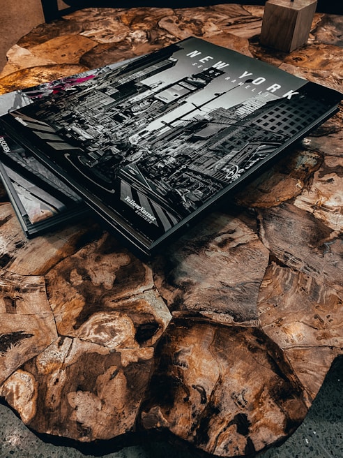 A wooden table with a unique, irregular pattern on its surface, featuring a large black-and-white photography book titled 'NEW YORK' depicting a city street scene. The book is partially overlaid with another book or magazine in the corner. A small wooden block is visible in the background.