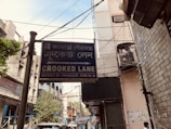 A street sign for 'Crooked Lane' appears prominently in the foreground, written in both English and Bengali. The street is flanked by buildings with visible utility wires overhead. The background reveals urban elements such as parked vehicles, trees, and signs of daily activity.