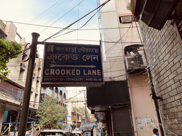 A street sign for 'Crooked Lane' appears prominently in the foreground, written in both English and Bengali. The street is flanked by buildings with visible utility wires overhead. The background reveals urban elements such as parked vehicles, trees, and signs of daily activity.