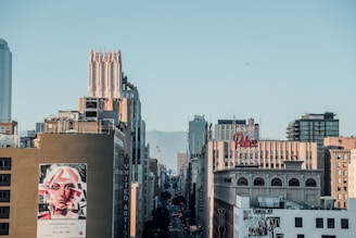 A bustling urban cityscape featuring a series of tall buildings along a main street. The architecture includes a mix of modern and older-style skyscrapers, with visible signage such as 'Palace Theatre' and 'United Artists'. A prominent billboard displays an artistic portrait of a woman. The background shows a clear sky with mountains faintly visible in the distance.