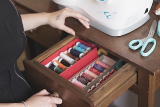 A pair of hands is next to a sewing machine with an open drawer containing multiple spools of thread in various colors. A pair of teal scissors is placed on the wooden table next to the machine.