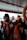 Inside a boat, several orange life jackets are neatly arranged on seats. A life preserver is visible on the right side. In the background, a person is looking down, possibly checking a mobile device, with hills and a structure visible through the open side of the boat.