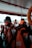 Inside a boat, several orange life jackets are neatly arranged on seats. A life preserver is visible on the right side. In the background, a person is looking down, possibly checking a mobile device, with hills and a structure visible through the open side of the boat.