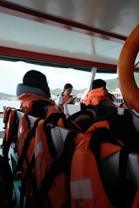 Inside a boat, several orange life jackets are neatly arranged on seats. A life preserver is visible on the right side. In the background, a person is looking down, possibly checking a mobile device, with hills and a structure visible through the open side of the boat.