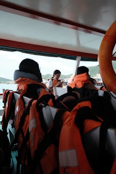 Inside a boat, several orange life jackets are neatly arranged on seats. A life preserver is visible on the right side. In the background, a person is looking down, possibly checking a mobile device, with hills and a structure visible through the open side of the boat.