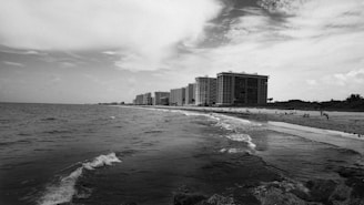 A black and white coastal scene featuring a series of tall apartment buildings lining the shore. The ocean spreads across the foreground with gentle waves touching the sandy beach. People are scattered along the shoreline and distant palm trees are visible near the buildings. The sky is partly cloudy, adding texture to the horizon.