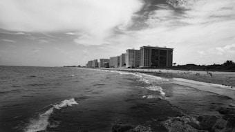 A black and white coastal scene featuring a series of tall apartment buildings lining the shore. The ocean spreads across the foreground with gentle waves touching the sandy beach. People are scattered along the shoreline and distant palm trees are visible near the buildings. The sky is partly cloudy, adding texture to the horizon.