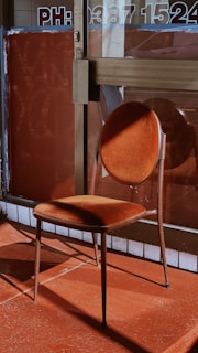 A vintage-style chair with a round backrest and square seat, both upholstered in a rich, velvet-like rust color. The chair is positioned in front of a glass entrance with a number and text partially visible on the door. The flooring and background share warm, earthy tones, creating a harmonious color palette with the chair.