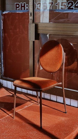A vintage-style chair with a round backrest and square seat, both upholstered in a rich, velvet-like rust color. The chair is positioned in front of a glass entrance with a number and text partially visible on the door. The flooring and background share warm, earthy tones, creating a harmonious color palette with the chair.