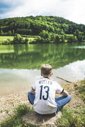 A person in a white sports jersey with the name Müller and number 13 is sitting by a lake. The individual faces away, gazing at the calm water with dense, green trees and a forested hillside in the background. The setting is serene and natural, with the sky partly cloudy.