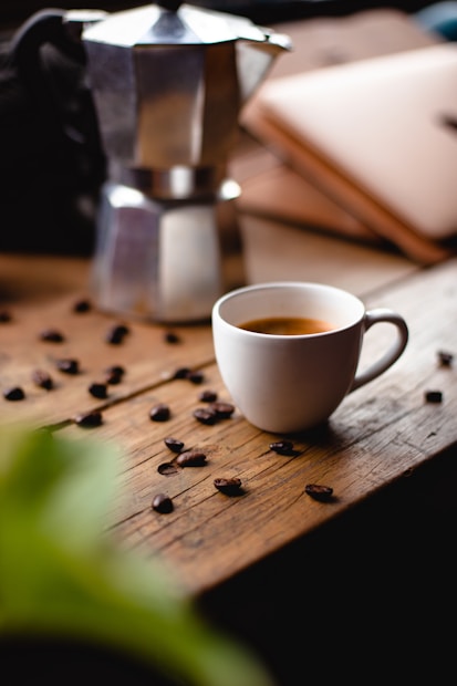 A white coffee cup filled with espresso is placed on a rustic wooden table. Scattered coffee beans surround the cup, adding to the cozy ambiance. A metal stovetop espresso maker sits in the background alongside what appears to be a closed laptop. Soft, natural lighting enhances the warm tones of the scene.
