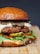 A gourmet hamburger with melted cheese, truffle slices, and a variety of toppings including pickles, arugula, and red bell pepper slices, all placed between a toasted bun. The presentation is on a wooden surface, showcasing a savory and tempting look.